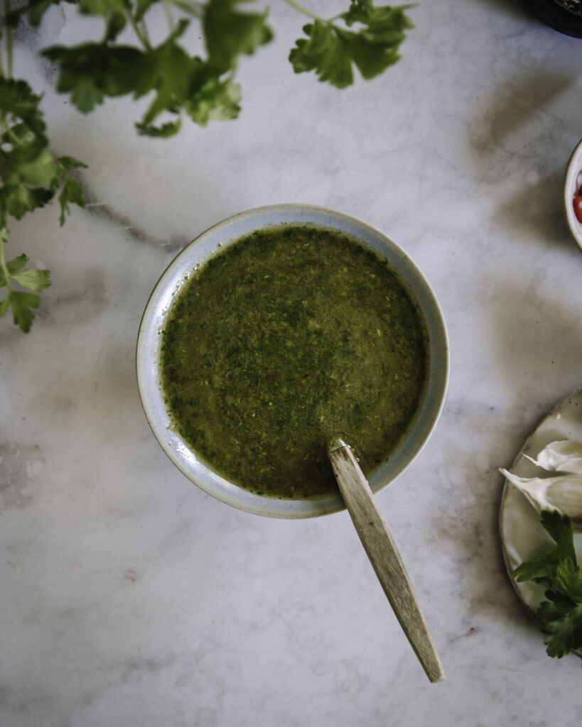 Chimichurri on helppo kastike, joka kruunaa monet ruoat. Kuvassa kastike on pyöreässä kulhossa. Kuva vasemmassa ylalaidassa on persiljanoksia ja oikeassa laidassa ainesosia lautasilla, joista näkyy vain osa.