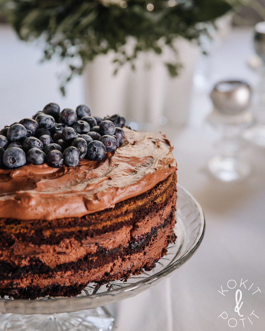 Täydellinen suklaamousse-täytekakku on lasisella kakkuvadilla. Taustalla näkyy sumeana juhlakattauksen kukat ja kynttilät.