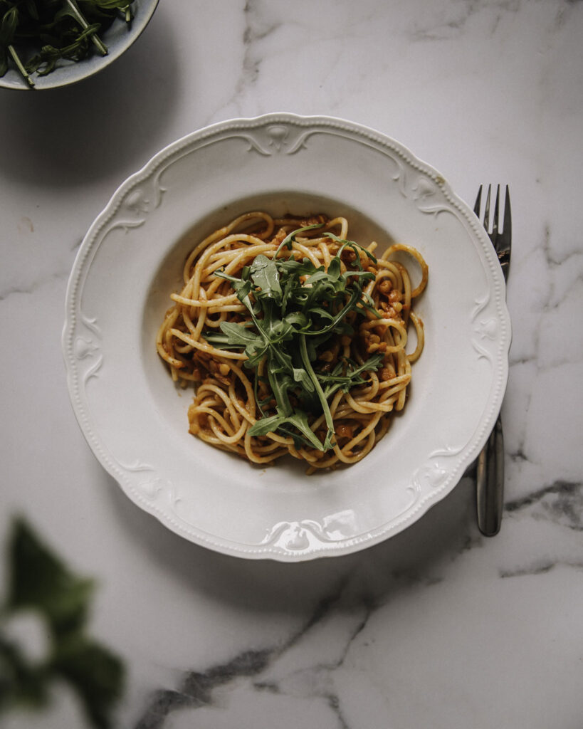 Linssibolognese on helppo ja nopea arkiruoka, joka syntyy kuiva-ainekaapin aineksista. Kuvassa kastike on sekoitettu nauhapastaan ja päällä on rucolaa. Pasta on valkoisella lautasella ja kuva on otettu ylhäältäpäin.