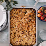 Soijarouhe-makaronilaatikko on helppo arkiruoka. Kuvassa makaronilaatikon ympärillä on lautasia, laseja ja kirsikkatomaatteja. Vasemmassa yläkulmassa on kukkia. Kuva alkusta on vaaleansininen.