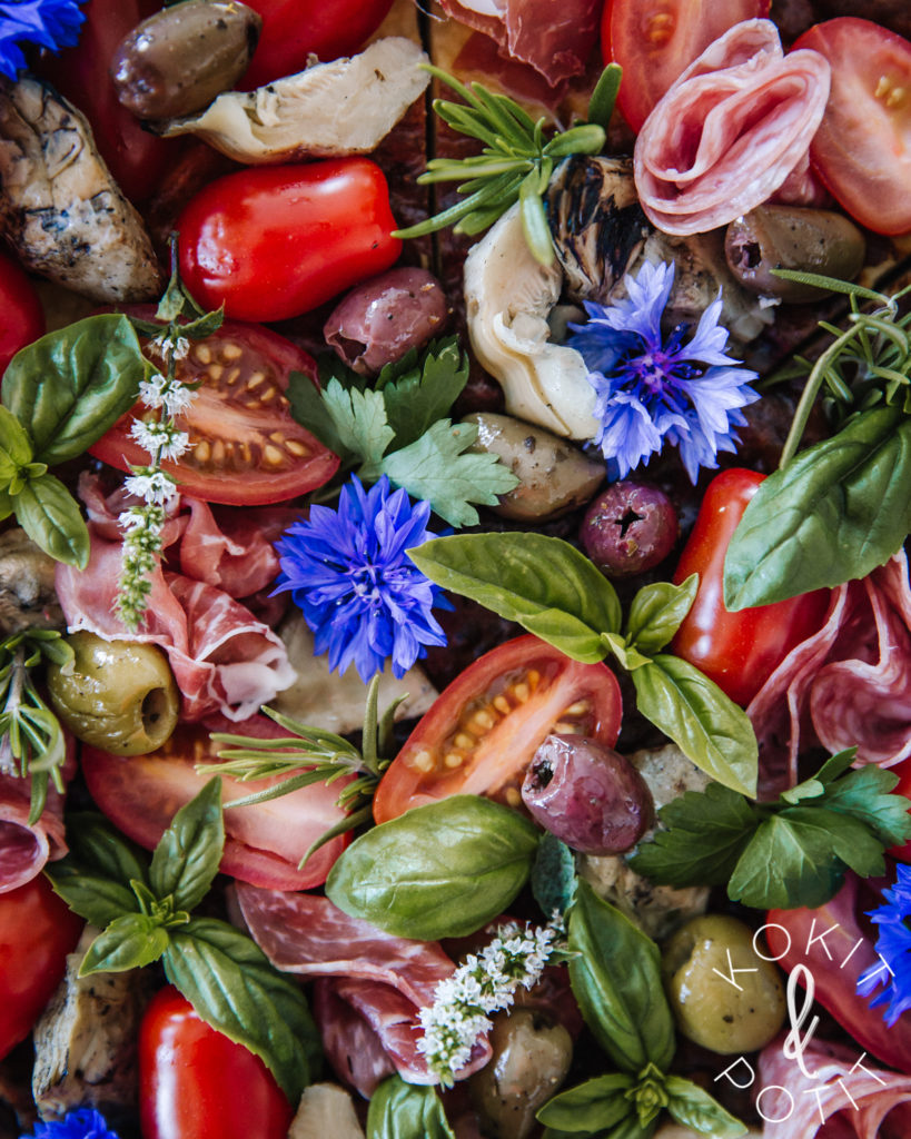 Kuva on täynnä tapaksia: oliiveja, erilaisia leikkeleitä, artisokansydämiä, yrttejä, syötäviä kukkia ja kirsikkatomaatteja.