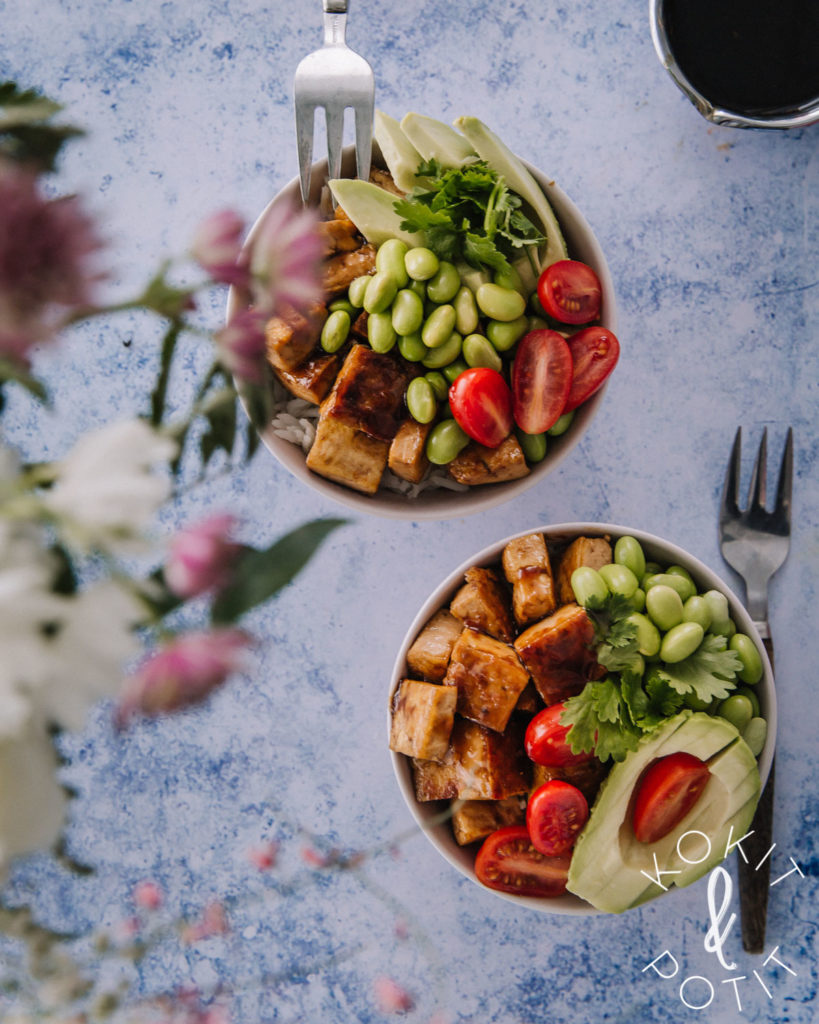 Vaaleansinisellä pöydällä on kaksi kulhoa, jossa on teriyaki-marinoitua tofua, papuja, avokadoa ja tomaattia. Alla on riisiä. Kuvan reunalla on kukkia, jotka ovat epätarkkoja.