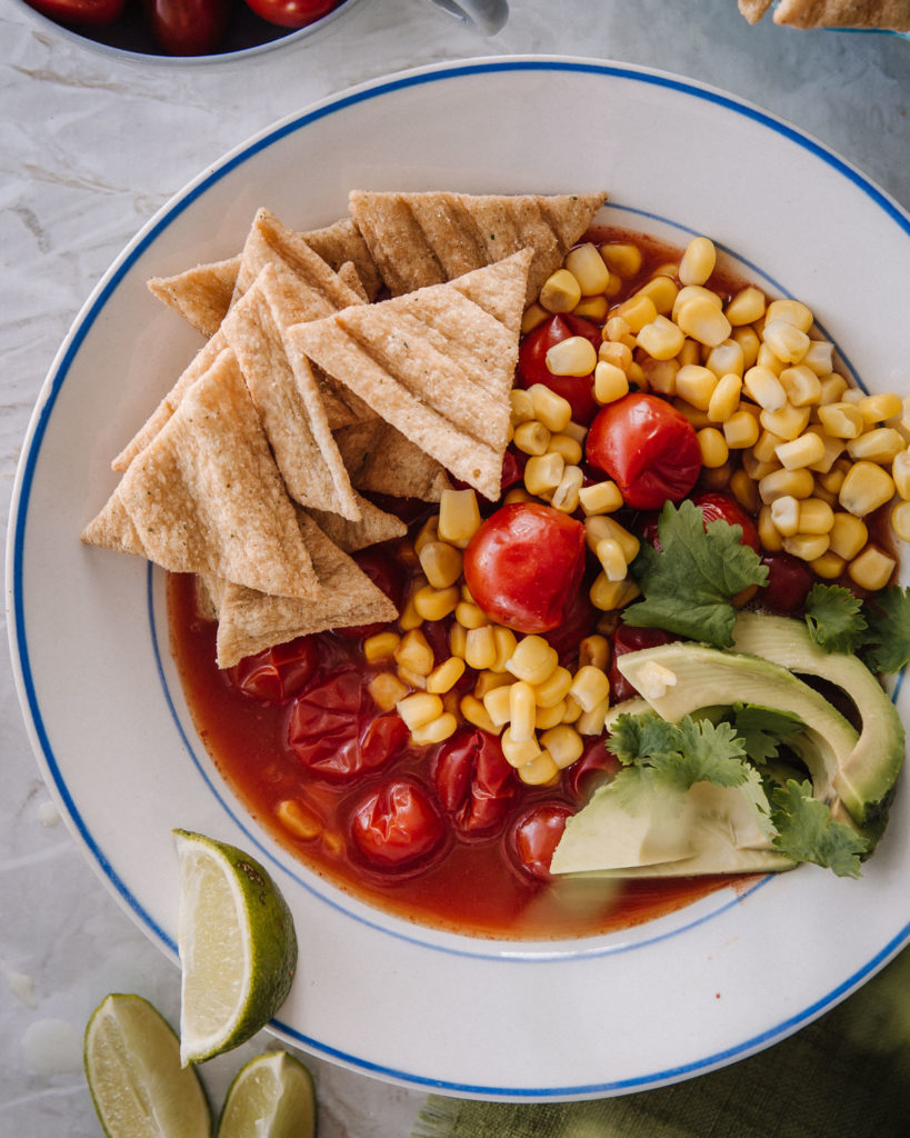 Meksikolainen tomaattikeitto on valkoisella lautasella. Päällä on avokadoa, kauranaksuja ja tuoretta korianteria. Keitossa on myös maissia.