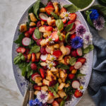 Mansikka-halloumisalaatti on sini-valkoisella, soikealla lautasella. Salaatin päällä on minttua, basilikaa sekä syötäviä kukkia. Kastike on pienessä kulhossa kuvan ylälaidassa.