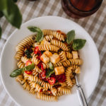 Halloumipasta on valkoisella syvällä lautasella. Kierrepasta, halloumikuutiot, uunissa paahdetut tomaatit ja basilikanlehdet ovat ihana yhdistelmä. Pöytäliina on ruudullinen.