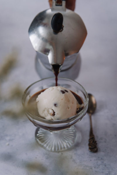 Affogato eli helppo italialainen jälkiruoka on laakeassa jalallisessa lasimaljassa. Kuvassa jäätelöpallon viereen kaadetaan espresso-kahvia. Lusikka on lasimaljan vieressä.