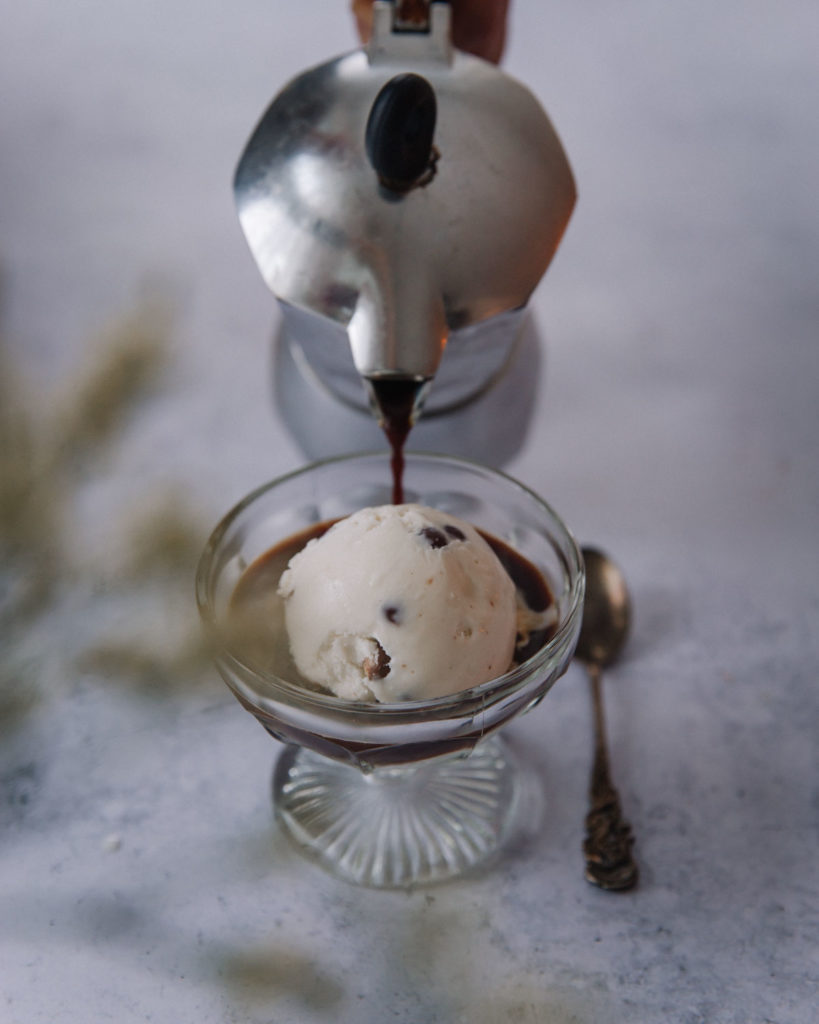 Affogato eli helppo italialainen jälkiruoka on laakeassa jalallisessa lasimaljassa. Kuvassa jäätelöpallon viereen kaadetaan espresso-kahvia. Lusikka on lasimaljan vieressä.