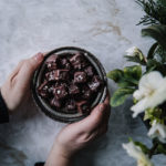 Maailman parhaat pipari-suklaakohvehdit on pyöreällä pienellä lautasella, jota kädet pitelevät. Oikeassa reunassa kuvaa on kukkia.
