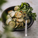 Kurkkusalaatti aasialaisittain on tummassa kahvallisessa keraamisessa astiassa. Salaatin sivussa on tuoretta korianteria. Korianteria on myös kuvan vasemmassa laidassa blurrina.