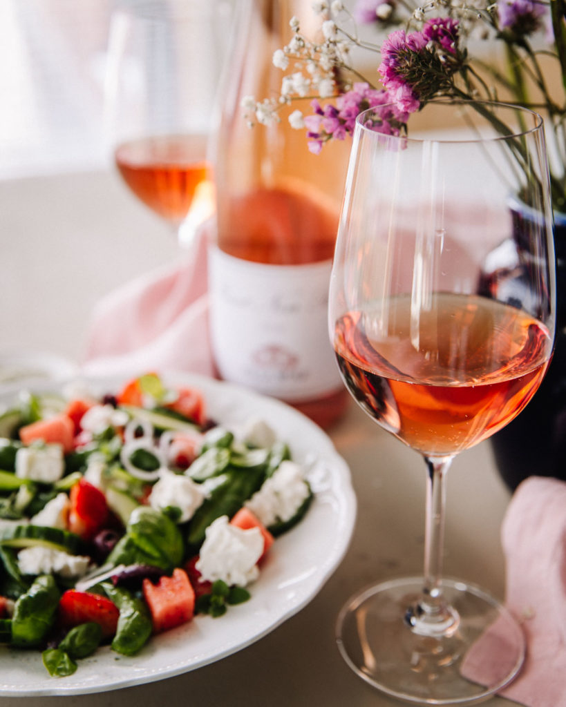Villa Wolf Pinot Noir Rosé on täydellinen valinta kesän kokkailuiden kaveriksi. Viinilasi on kuvan oikeassa reunassa. Kuvassa on lisäksi epätarkkana kesäinen salaatti, viinipuollo sekä kukkia.