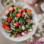 Kreikkalainen salaatti mansikoilla ja vesimelonilla. Salaatti on valkoisella lautasella, jonka reunassa on kohokuvioita. Kuva on ilmeeltään runsas. Reunoilla on kukkia, viinilaseja ja serviettejä.