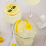 Limoncello spritz on täydellinen kesädrinkki! Kuvassa on kolme erilaista lasia. Kahdessa lasissa on koristeena sitruunaviipale. Kuvan oikeassa laidassa on iso jääpala ja vasemmassa alakulmassa pari keltaista kukkaa.