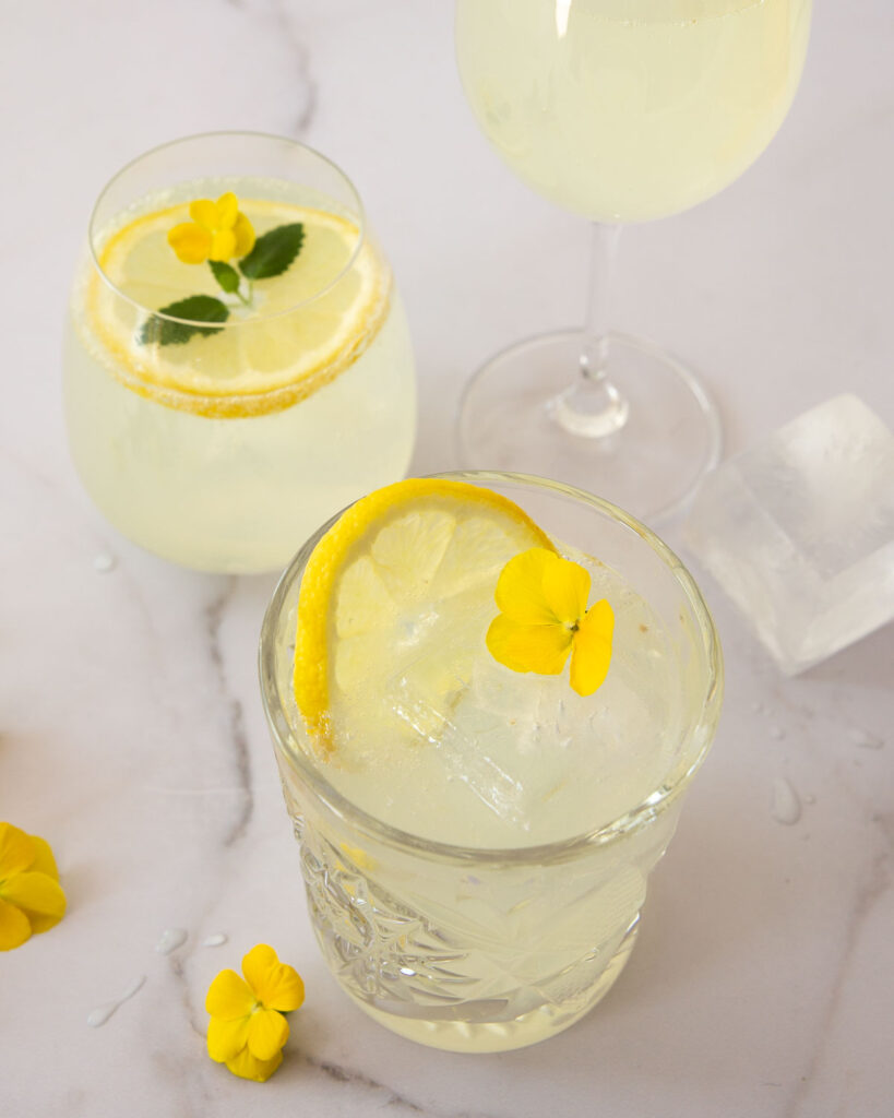 Limoncello spritz on täydellinen kesädrinkki! Kuvassa on kolme erilaista lasia. Kahdessa lasissa on koristeena sitruunaviipale. Kuvan oikeassa laidassa on iso jääpala ja vasemmassa alakulmassa pari keltaista kukkaa.