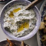 Tzatziki eli kreikkalainen jogurttikastike. Kastike on sinivalkoisessa kulhossa ja kastikkeen päällä on mustapippuria rouheena sekä loraus oliiviöljyä. Kuva on otettu ylhäältäpäin.
