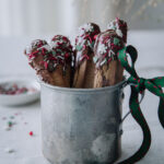 Helpot piparitikut suklaakuorrutteella on kuvassa peltimukissa pystyssä. Tikkujen päät on kastettu suklaaseen ja suklaan päälle on ripoteltu joulu-kuvioisia nonparelleja. Mukin korvassa on vihreäpunainen rusetti.
