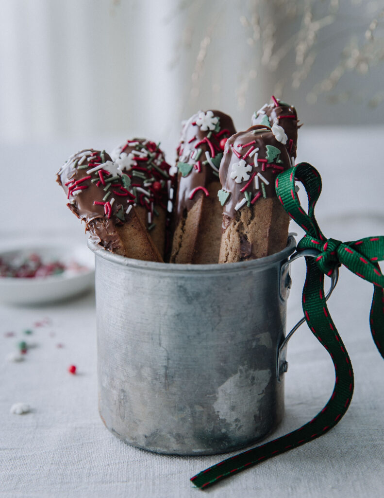 Helpot piparitikut suklaakuorrutteella on kuvassa peltimukissa pystyssä. Tikkujen päät on kastettu suklaaseen ja suklaan päälle on ripoteltu joulu-kuvioisia nonparelleja. Mukin korvassa on vihreäpunainen rusetti.