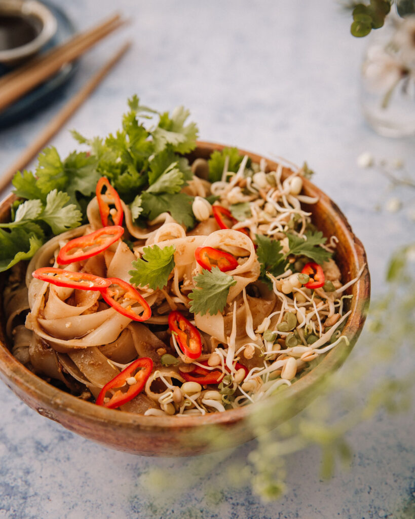 Pad Thai eli thaimaalaiset paistetut nuudelit – yhtä hyvää kuin Thaimaassa. 

Kuvassa ruoka on keraamisessa kulhossa. Päällä on punaisia chiliviipaleita, korianteria ja ituja. Kuvan etualalla on epätarkkoja kasveja. kuva on otettu 45-asteen kulmassa.