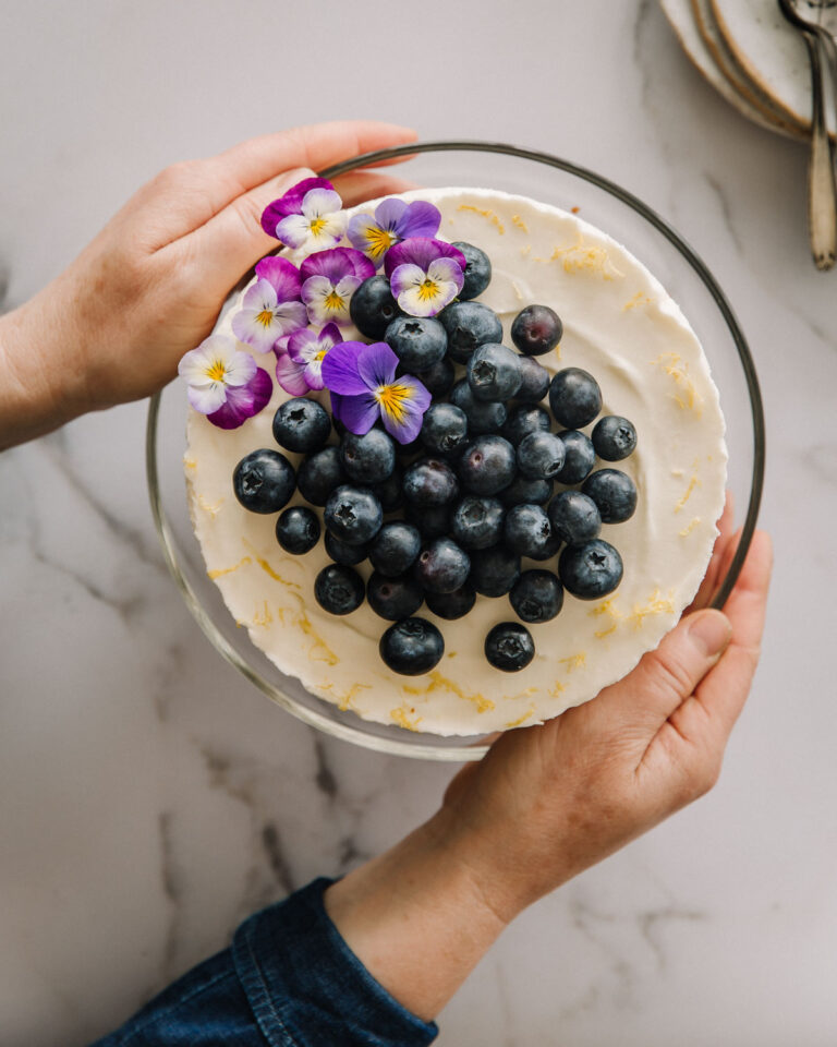 Sitruunajuustokakku on ihana juhlien suosikki kakku! Kakku on jalallisella kakkulautasella, kädet ovat lautasen molemmin puolin. Kakun päällä on pensasmustikoita ja orvokkeja, kakun reunoilla on sitruunan kuorta raastettuna. Kuva on otettu ylhäältäpäin.