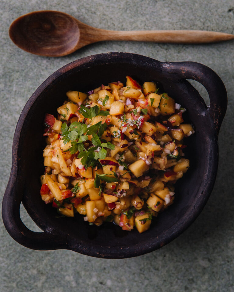 Nektariinisalsa on helppo ja herkullinen lisäke. Kuvassa salsa on tummassa, pyöreässä astiassa, jossa on pienet kahvat. Salsan päällä on korianteria. Astian yläpuolella on puinen lusikka.