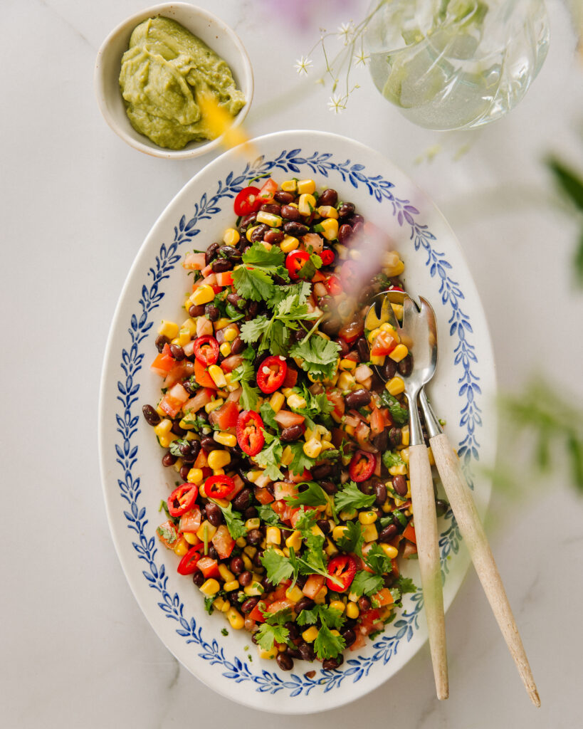 Papusalsa on helppo tehdä. Kuvassa salsa on soikealla tarjoilulautasella. Lautasella on myös ottimet. Oikeassa reunassa ja ylhäällä on epätarkkana kasveja. Klo 11 kohdalla on pienessä kipossa avokadotahnaa. Kuva on otettu ylhäältäpäin.