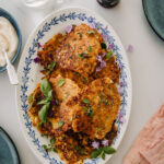 Linssi-kasvispihvit ovat soikealla tarjoiluvadilla, joka on valkoinen sinisellä reunakuviolla. Pihvit ovat oranssin sävyisiä. Pihvien päällä on hieman yrttejä sekä syötäviä kukkia. Kuvan reunoilla on muita astioita osin näkyvissä. Kuva on otetu ylhäältäpäin.