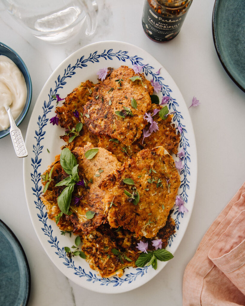 Linssi-kasvispihvit ovat soikealla tarjoiluvadilla, joka on valkoinen sinisellä reunakuviolla. Pihvit ovat oranssin sävyisiä. Pihvien päällä on hieman yrttejä sekä syötäviä kukkia. Kuvan reunoilla on muita astioita osin näkyvissä. Kuva on otetu ylhäältäpäin.