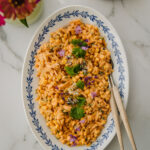 Maissi-pastasalaatti on kullankeltainen herkku. Pasta on pientä orzoa. Salaatti on soikealla vadilla, jonka reunoilla on sininen lehväkuvio. Salaatin päällä on korianteria ja syötäviä kukkia. Kuva yläreunassa on kukkia ja lautasia. Kuva on otettu ylhäältäpäin.