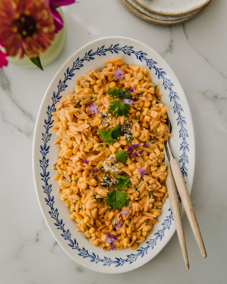 Maissi-pastasalaatti on kullankeltainen herkku. Pasta on pientä orzoa. Salaatti on soikealla vadilla, jonka reunoilla on sininen lehväkuvio. Salaatin päällä on korianteria ja syötäviä kukkia. Kuva yläreunassa on kukkia ja lautasia. Kuva on otettu ylhäältäpäin.