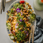 Ruokaisa salaatti seitanilla on moneen tilanteeseen sopiva ruoka. Salaatti on vegaaninen. Kuvassa salaatti on soikealla vadilla. Päällimmäisenä on yrttejä ja syötäviä kukkia. Kuva ylälaidassa on lautaspino sekä haarukoita sekä osa kukkavaasista. Oikeassa alakulmassa on kankainen servetti. Kuva on otettu ylhäältäpäin.