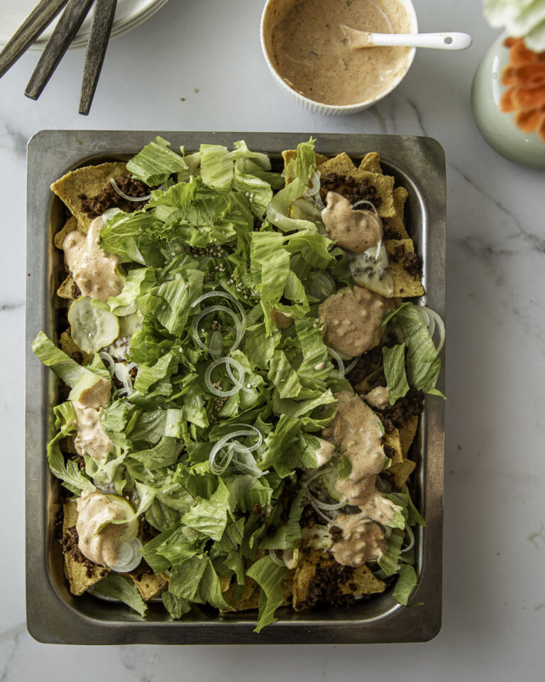 Big Mac -nachopelti hurmaa. Nachojen päällä on hampurilaisesta tuttuja makuja. Päällimmäisenä on rapeaa salaattia ja sipulirenkaita sekä kastiketta. Kuva ylälaidassa on lautasia ja haarukoita sekä kukkia.