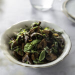 Marinoidut herkkusienet on monikäyttöinen lisäke tai pikkuruoka. Sienet ovat pyörässä kulhossa, jonka reunat ovat aaltoilevat ja reunassa on ohut kultainen viiva. Sieniä päällä on tuoretta basilikaa. Kuva on vaalea ja sen yläosassa on pieniä lautasia sekä osa vesikannua.