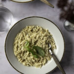 Sitruunainen orzo - yhden astian pasta on helppo arkiruoka. Pasta on valkoisella, kultareunaisella lautasella. Pastan päällä on basilikaa ja kello viidessä on puuvartinen haarukka. Kuvan yläosassa näkyy toisen lautasen reuna ja tummaa kukkaa.