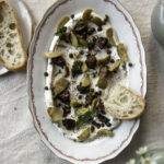 Vuohen juustodippi on yhdistelmä dippiä ja tapaksia. Dippi on laakealla vadilla, jossa on punaista kuviota reunoilla. Vaalean dipin päällä on paloina oliiveja, sieniä sekä pieniä kapriksia ja yrttejä. Kuvan oikeassa laidassa on vaaleaa leipää.