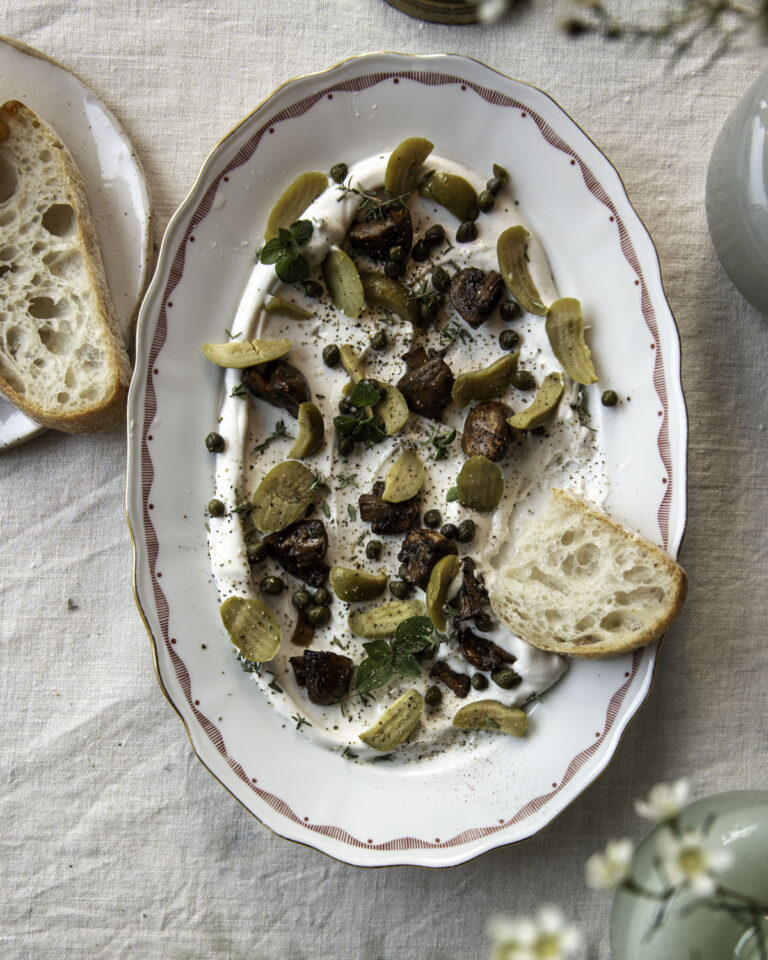 Vuohen juustodippi on yhdistelmä dippiä ja tapaksia. Dippi on laakealla vadilla, jossa on punaista kuviota reunoilla. Vaalean dipin päällä on paloina oliiveja, sieniä sekä pieniä kapriksia ja yrttejä. Kuvan oikeassa laidassa on vaaleaa leipää.