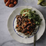 Chili sin carne on helppo vegaaninen pataruoka. Kuvassa ruoka on vaalealla lautasella riisin päällä. Lautasella on myös korianteria ja avokadoa. Päällä on punaisia chilirenkaita. Kuva yläreunassa on limenlohkoja, juomalasi ja kirsikkatomaatteja.