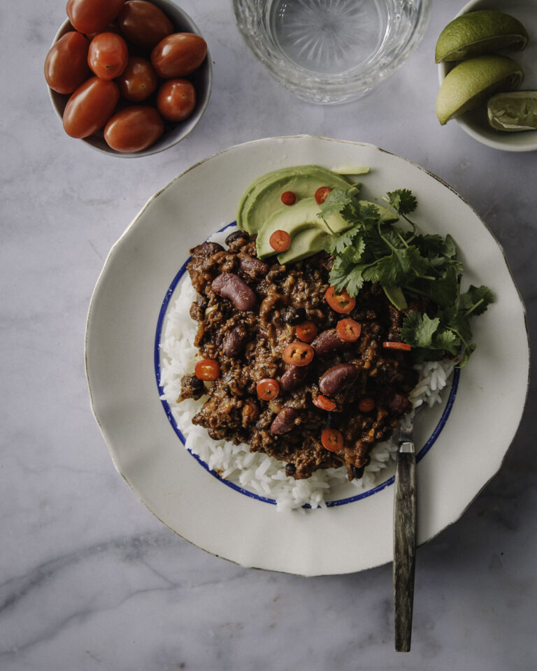 Chili sin carne on helppo vegaaninen pataruoka. Kuvassa ruoka on vaalealla lautasella riisin päällä. Lautasella on myös korianteria ja avokadoa. Päällä on punaisia chilirenkaita. Kuva yläreunassa on limenlohkoja, juomalasi ja kirsikkatomaatteja.