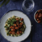 Turkkilainen pasta on somen villinnyt hittiruoka. Kuvassa ruoka on valkoisella lautasella, jonka reunassa on kultainen reuna. Pastan päällä on puolitettuja kirsikkatomaatteja sekä lehtipersiljaa. Kuvan tausta on sininen.