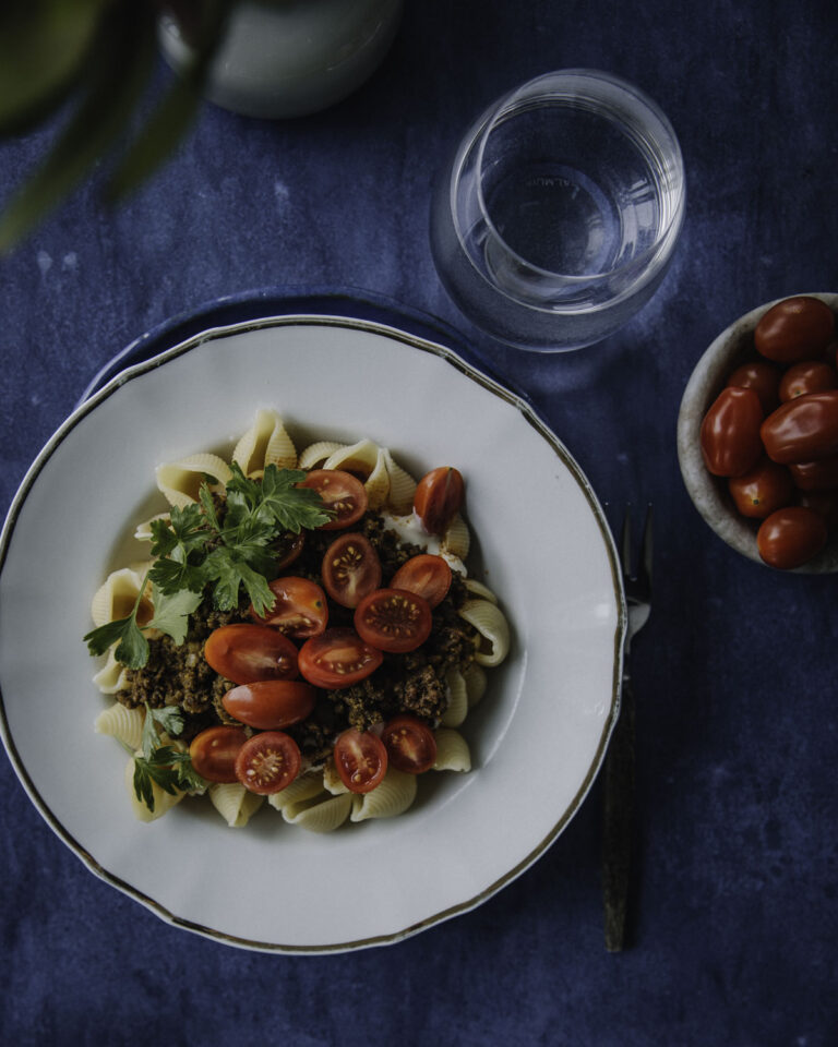 Turkkilainen pasta on somen villinnyt hittiruoka. Kuvassa ruoka on valkoisella lautasella, jonka reunassa on kultainen reuna. Pastan päällä on puolitettuja kirsikkatomaatteja sekä lehtipersiljaa. Kuvan tausta on sininen.