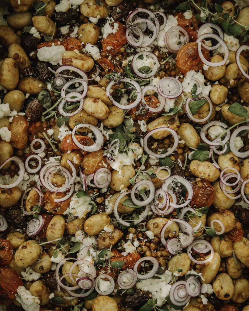 Helppo gnocchivuoka Välimeren mauilla on maukas arkiruoka. Kuva on lähikuva ruoasta. Gnocchien joukossa on linssejä, oliiveja ja uunissa pehmenneitä kirsikkatomaatteja. Ruoan päällä on yrttejä ja punasipulirenkaita.