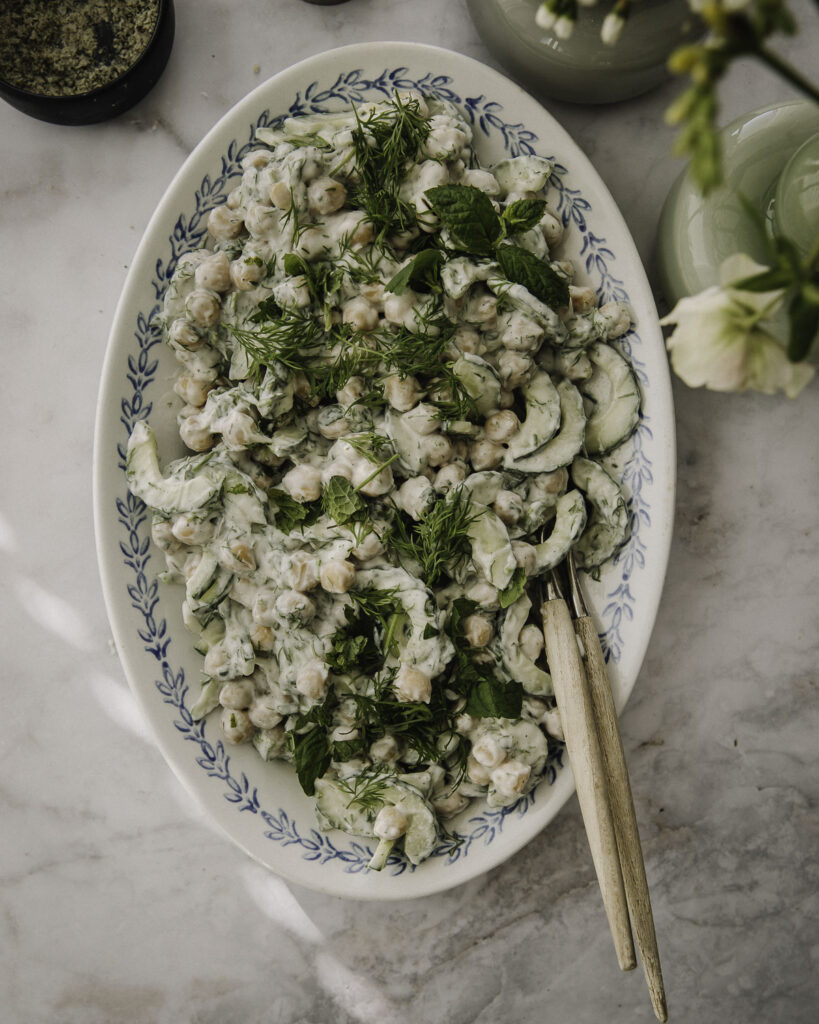 Tzatziki-kikhernesalaatti on helppo ja herkullinen lisäke. Kuvassa salaatti on vaalealla soikealla lautasella, jossa on sininen kuvio reunalla. Salaatin päällä on tilliä ja mintunlehtiä.