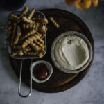 Aioli eli valkosipulimajoneesi on helppo tehdä itse. Kuvassa aioli on pyöreässä pienessä kupissa. Vieressä on kahvallisessa teräskorissa ranskalaisia. Pyöreän puualustan päällä on myös pienessä astiassa ketsuppia.