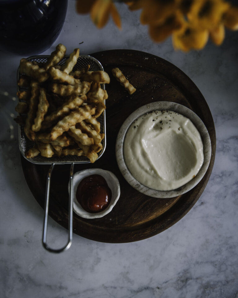 Aioli eli valkosipulimajoneesi on helppo tehdä itse. Kuvassa aioli on pyöreässä pienessä kupissa. Vieressä on kahvallisessa teräskorissa ranskalaisia. Pyöreän puualustan päällä on myös pienessä astiassa ketsuppia.
