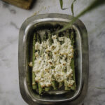 Paahdetut parsat valmistuvat uunissa vartissa. Kuvassa parsat ovat metallisella vadilla ja parsojen päällä on feta-sitruunakuorrute. Kuvan ylälaidessa on kasvin lehtiä sekä leikkuulauta, jolla on puolikas sitruuna. Kuva on otettu ylhäältäpäin.