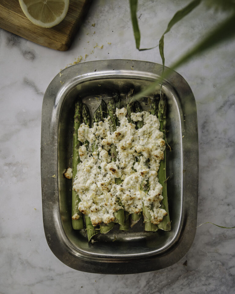 Paahdetut parsat valmistuvat uunissa vartissa. Kuvassa parsat ovat metallisella vadilla ja parsojen päällä on feta-sitruunakuorrute. Kuvan ylälaidessa on kasvin lehtiä sekä leikkuulauta, jolla on puolikas sitruuna. Kuva on otettu ylhäältäpäin.