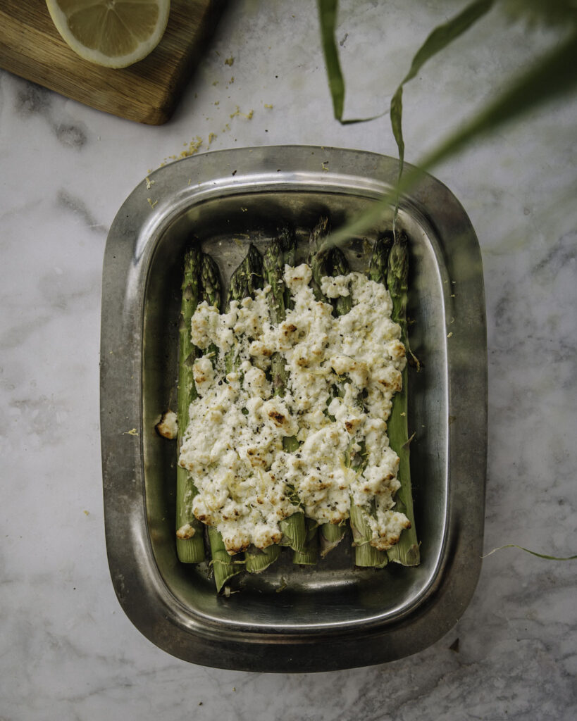 Paahdetut parsat valmistuvat uunissa vartissa. Kuvassa parsat ovat metallisella vadilla ja parsojen päällä on feta-sitruunakuorrute. Kuvan ylälaidessa on kasvin lehtiä sekä leikkuulauta, jolla on puolikas sitruuna. Kuva on otettu ylhäältäpäin.