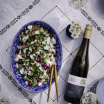 Feta-perunasalaatti on helppo ja herkullinen kesäruoka. Kuvassa ruoka on sinisellä vadilla. Ruoan päällä on fetaa, herneitä ja syötäviä kukkia. Salaatin vieressä on valkoviinipullo.