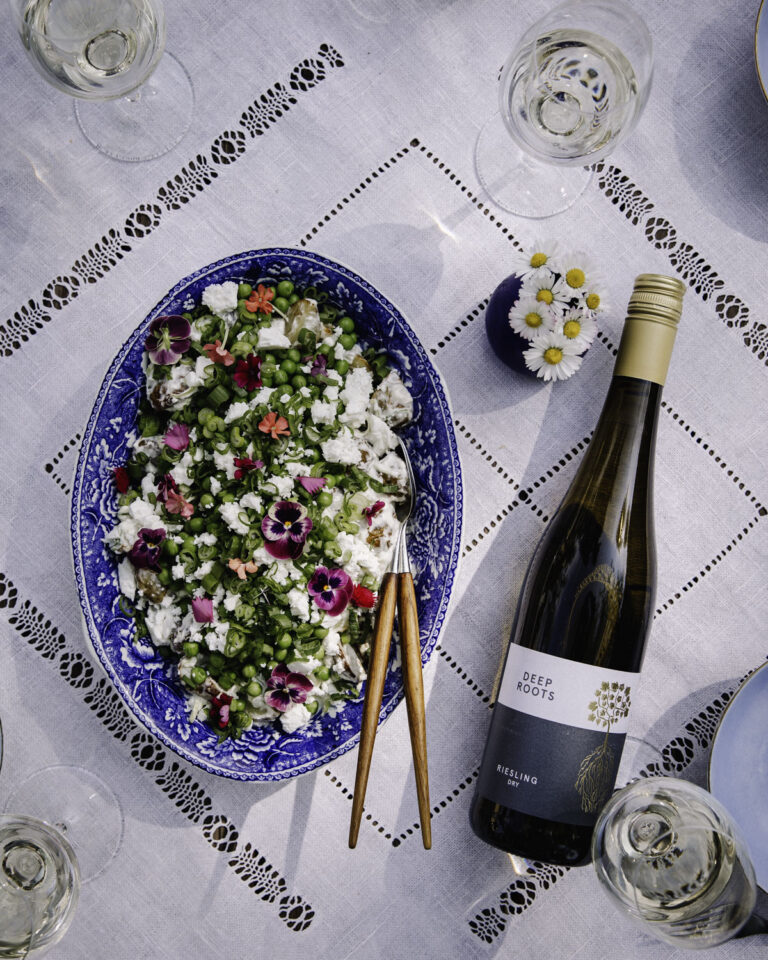 Feta-perunasalaatti on helppo ja herkullinen kesäruoka. Kuvassa ruoka on sinisellä vadilla. Ruoan päällä on fetaa, herneitä ja syötäviä kukkia. Salaatin vieressä on valkoviinipullo.