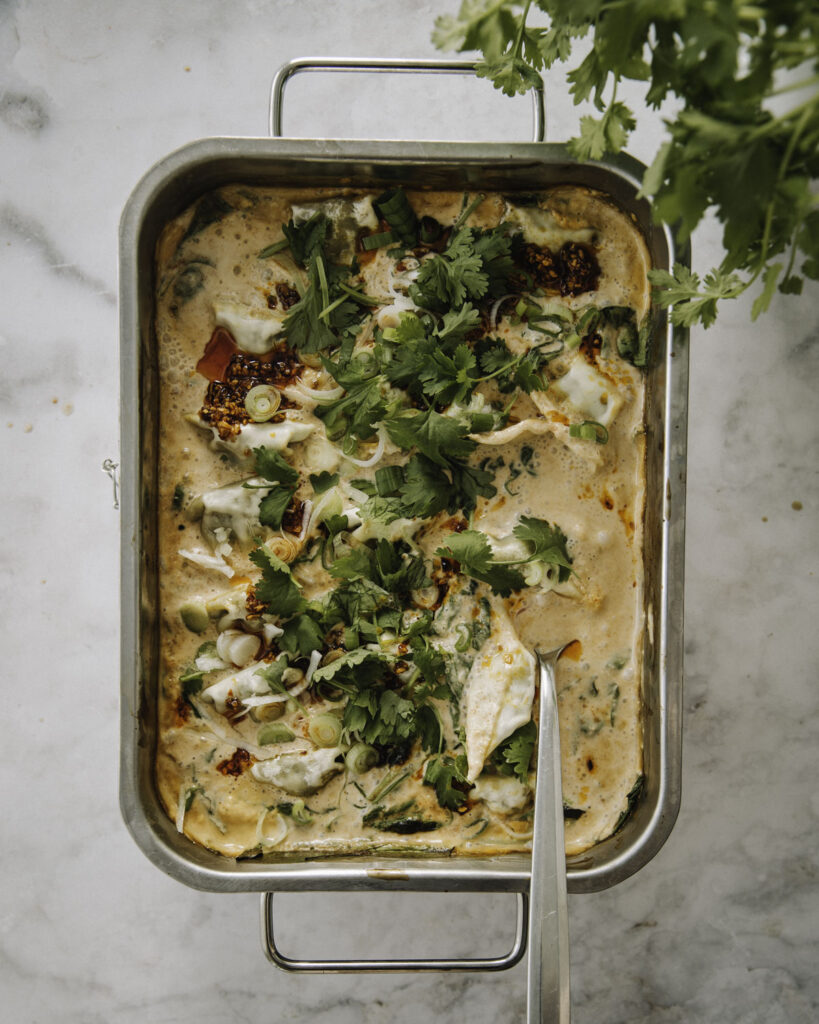 Helppo dumpling-paistos valmistuu alle puolessa tunnissa uunissa. Kuvassa ruoka on suorakulmaisessa uunivuoassa. Ruoan päällä on kesäsipulia, korianteria ja rapeaa valkosipuli-chiliöljyä. Kuvan oikeassa yläkulmassa on korianteripuska.