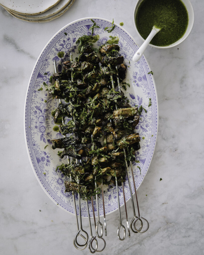 Sienivartaat on helppo grilliruoka. Kuvassa vartaat on metallisissa tuplatikuissa. Grillattujen vartaiden päällä on vihreää chimichurri-kastiketta ja yrttisilppua. Vartaat ovat soikealla vadilla.