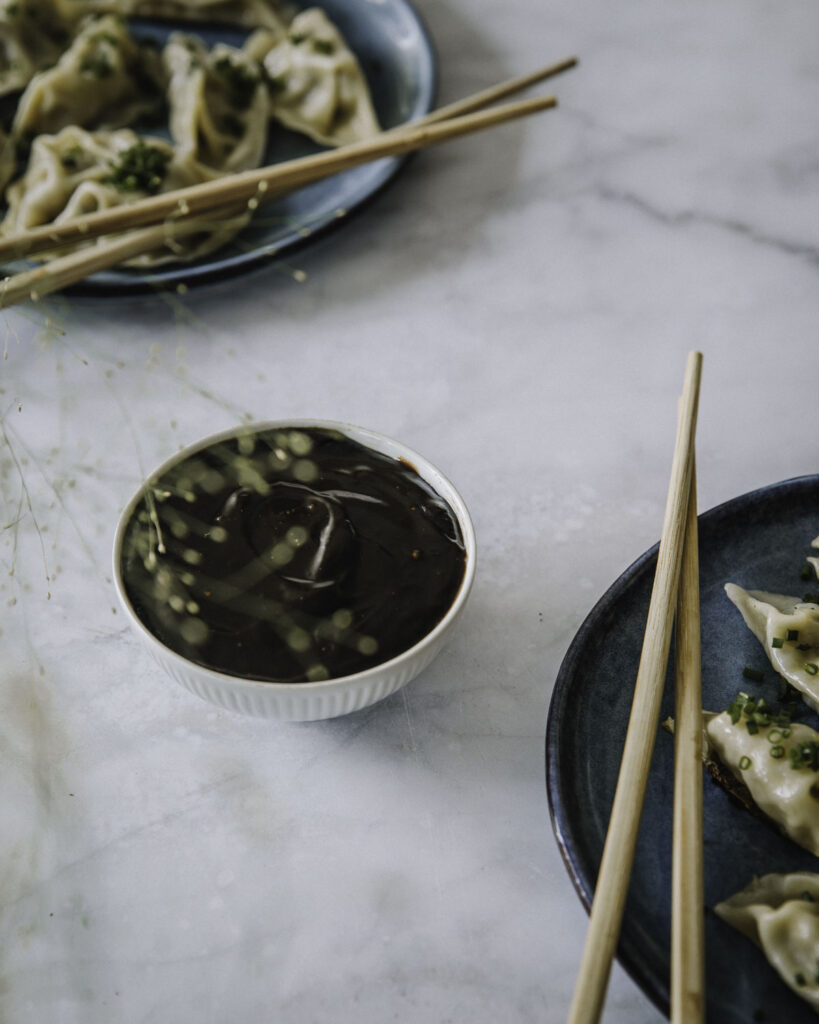 Hoisin-kastike on aasialainen kastike, joka sopii dipiksi, pöytämausteeksi tai vaikka wokkien viimeistelyyn. Kuvassa kastike on pienessä valkoisessa kulhossa. Kuvan oikeassa alalaidassa ja vasemmassa yläkulmassa on lautaset, joilla on dumpingeja.